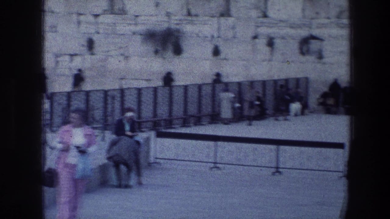 1976 8mm frame of visitors near the Wailing Wall area in Jerusalem, with people walking across the courtyard and others seated or standing near the stone wall.