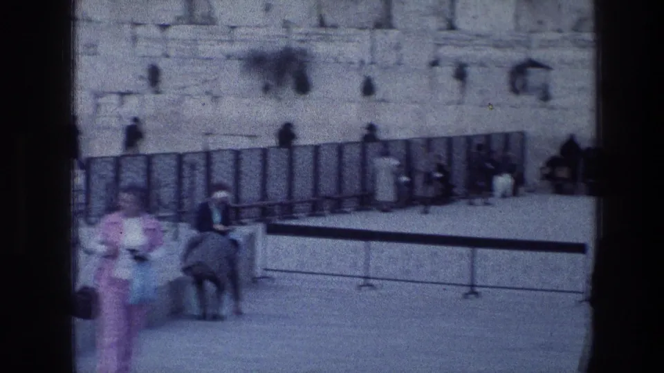 1976 8mm frame of visitors near the Wailing Wall area in Jerusalem, with people walking across the courtyard and others seated or standing near the stone wall.