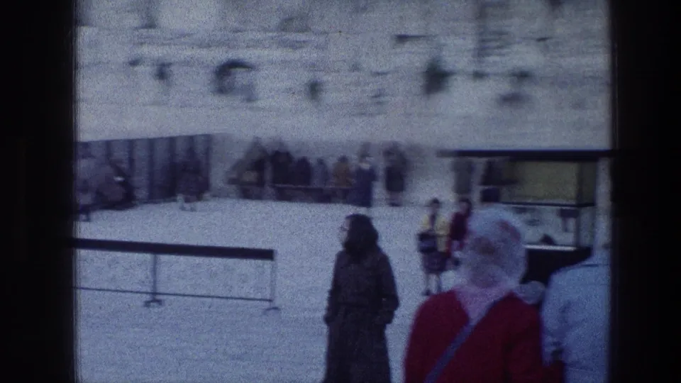 1976 home-movie frame near the Wailing Wall in Jerusalem showing visitors in coats and a woman in red moving through the stone courtyard.