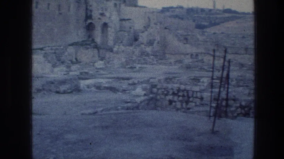 1976 home-movie frame in Jerusalem showing rocky ground, low stonework, metal bars, and weathered stone structures near the Wailing Wall surroundings.