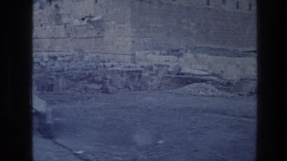 Blue-tinted 1976 home-movie frame showing the lower Wailing Wall area, scattered stones, and open ground without visible visitors.