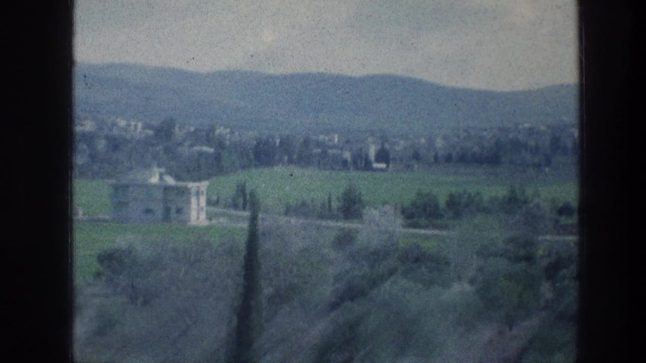 Green countryside with a pale villa, trees, distant hills, and dark film borders.
