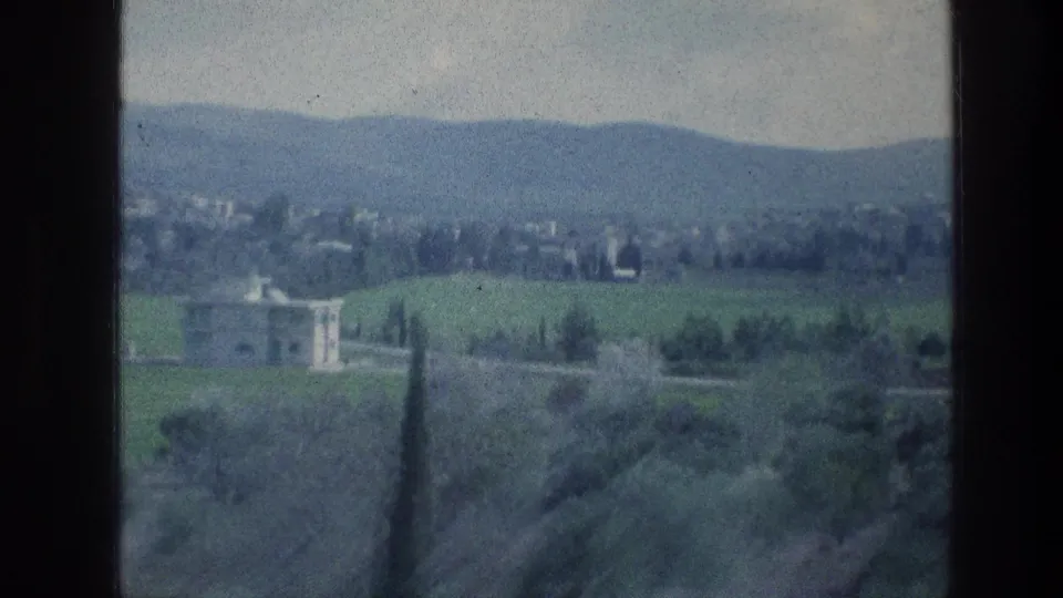 1976 8mm frame of a green hillside landscape near the stated Jerusalem-area reel, with a pale villa-like building and distant hills.