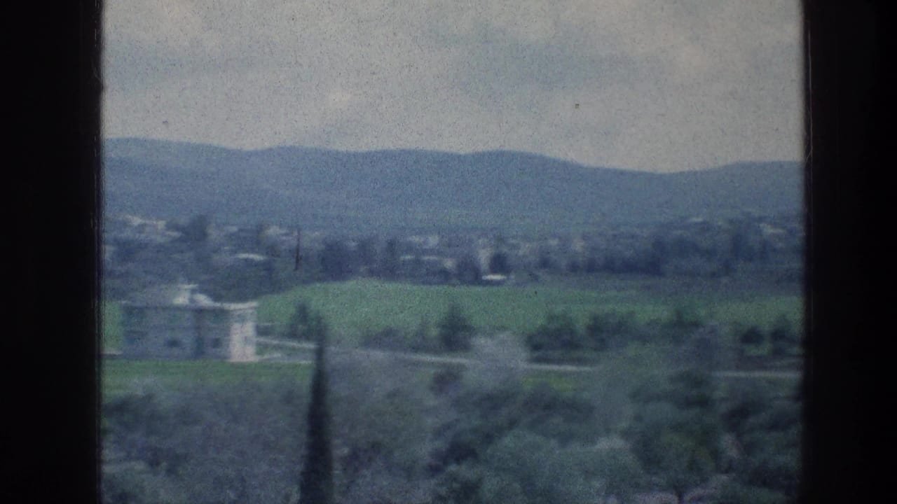 Countryside view with green fields, trees, a pale building, and low hills in the distance.