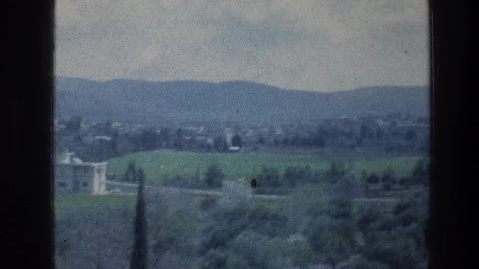1970s 8mm frame of a wide rural landscape with green fields, a pale building at left, scattered trees, and hazy hills.