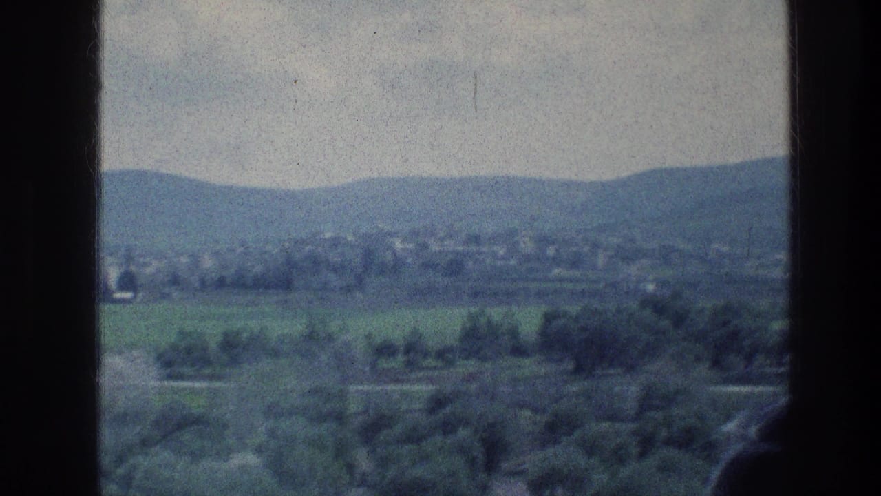 1976 8mm view near Jerusalem showing green hills, distant settlement, cloudy sky, and dark foreground window or vehicle edges.