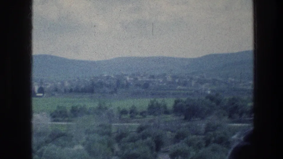 1976 8mm view near Jerusalem showing green hills, distant settlement, cloudy sky, and dark foreground window or vehicle edges.