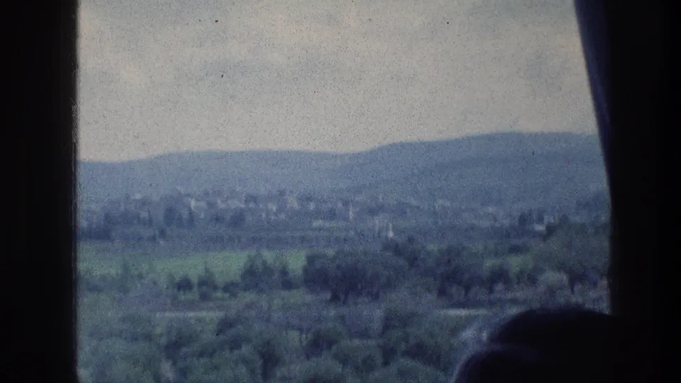 1976 8mm travel frame near Jerusalem with a green valley, low hills, distant structures, and dark window-like foreground edges.