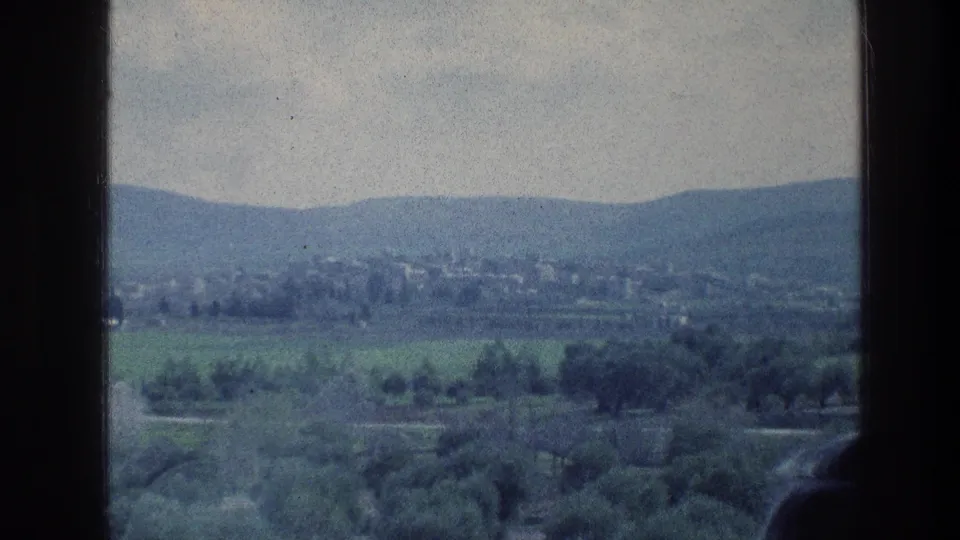 1976 8mm view near Jerusalem with green fields, tree rows, distant buildings, hills, cloudy sky, and heavy dark foreground framing.
