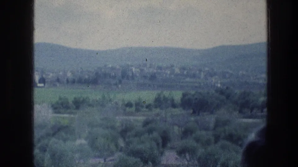 1976 8mm Jerusalem-area landscape frame showing hills, green tree cover, distant settlement, cloudy sky, and a partial foreground figure edge.