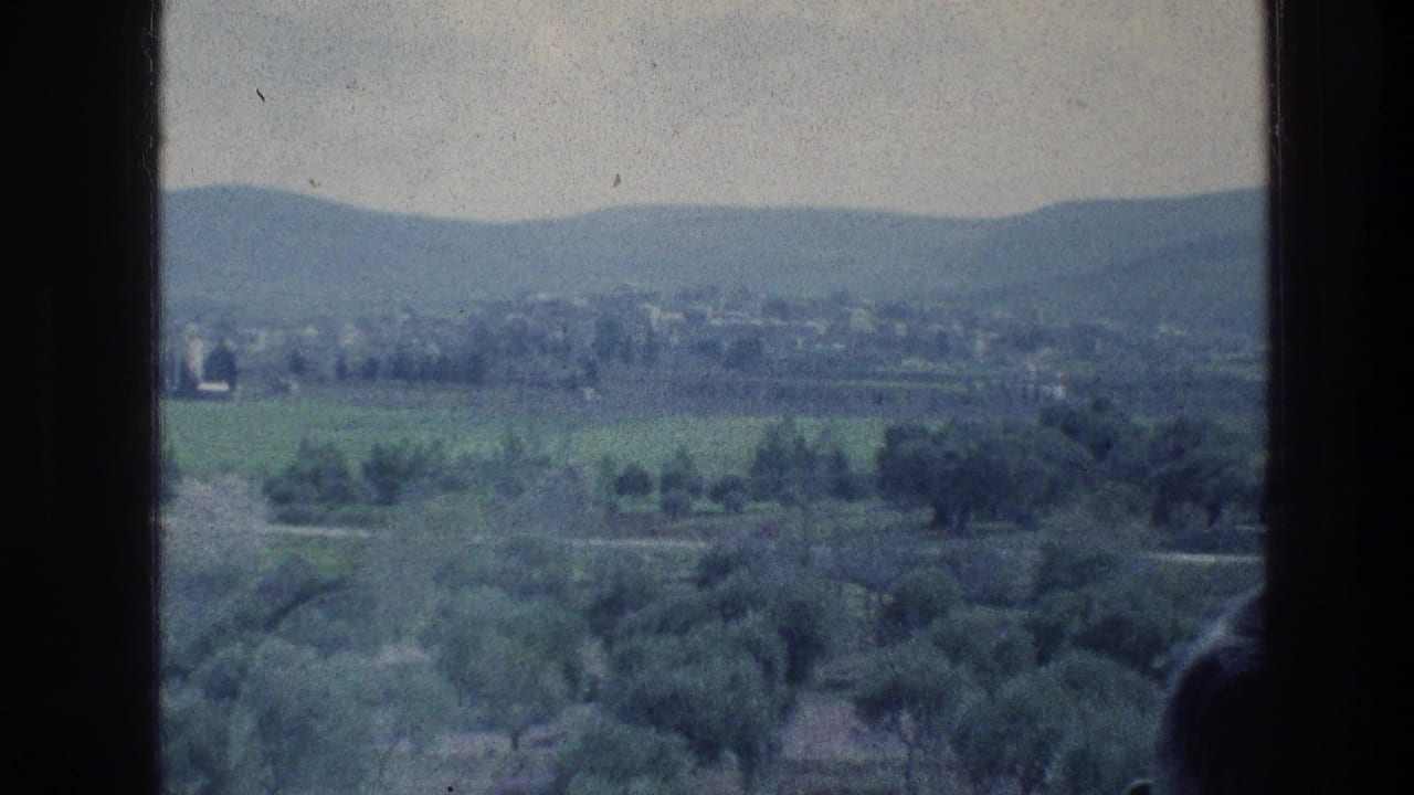 1976 8mm-style view of green fields, trees, distant buildings, and low hills framed by dark vertical edges.
