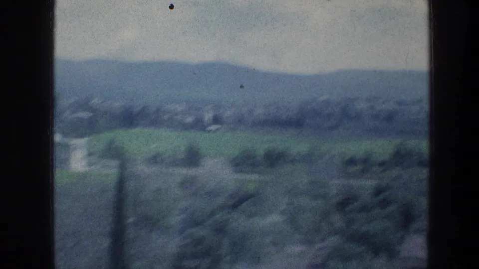 Muted 8mm frame showing a hazy rural panorama with foreground blur, fields, tree cover, and distant hills.