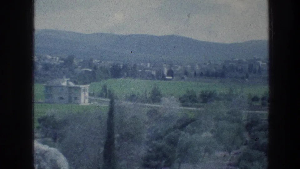 1976 8mm home-movie frame with a pale building, green fields, trees, and hazy hills in the distance.