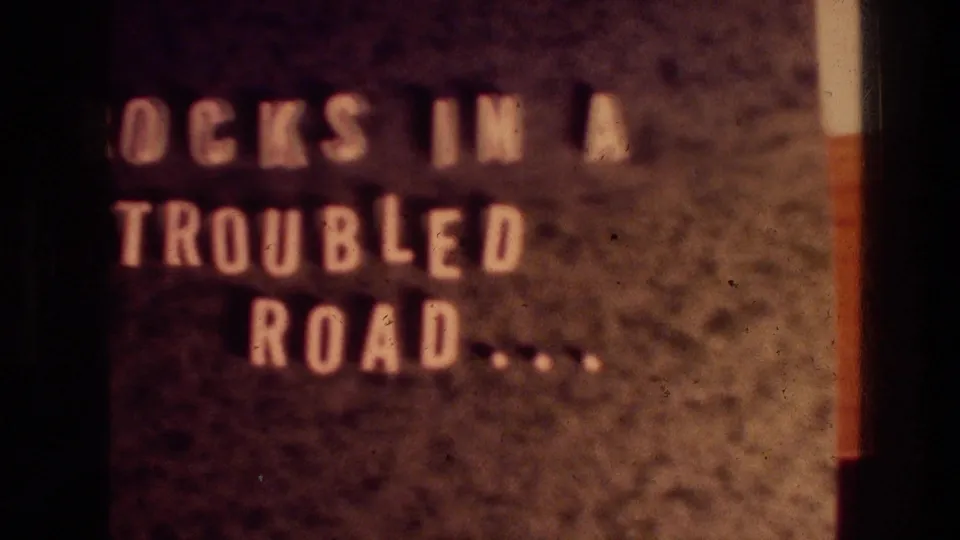 1976 8mm title-card frame with cropped pale lettering reading part of "ROCKS IN A TROUBLED ROAD" on a dark background.