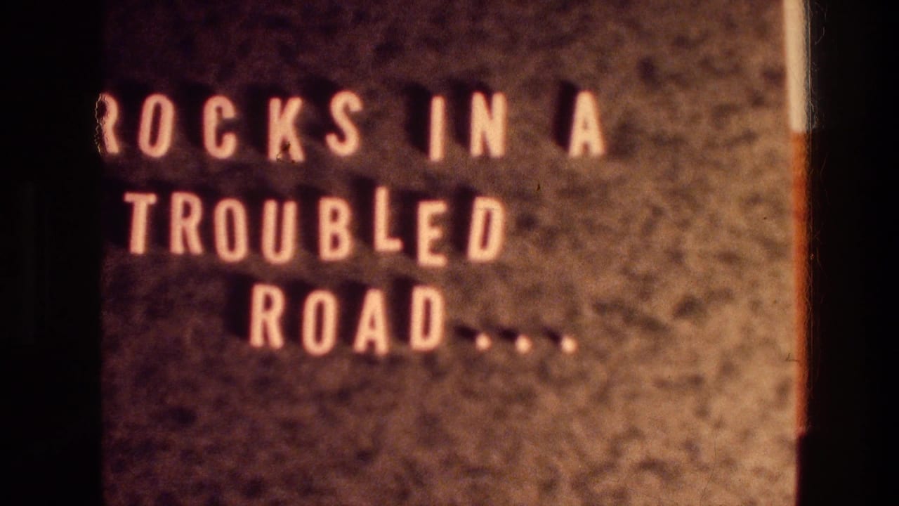 1976 8mm frame showing a title card with pale block lettering, "ROCKS IN A TROUBLED ROAD.....," against a dark textured field.