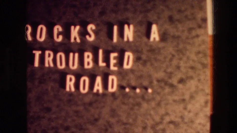 1976 8mm frame showing a title card with pale block lettering, "ROCKS IN A TROUBLED ROAD.....," against a dark textured field.