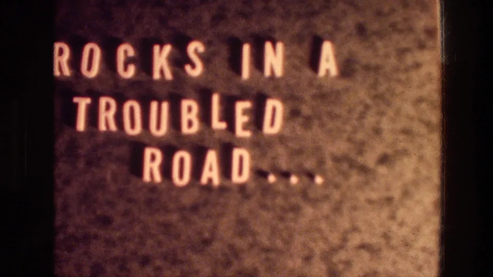 1976 home-movie title-card frame, cropped close on the words "ROCKS IN A TROUBLED ROAD" with visible grain and gate edges.