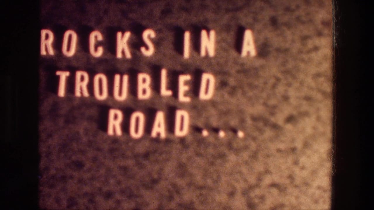 1976 8mm title card with pale letters reading ROCKS IN A TROUBLED ROAD on a warm textured background.