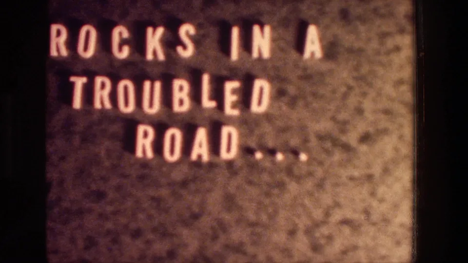 Close 8mm view of the title card ROCKS IN A TROUBLED ROAD, cropped by the film frame and softened by grain.