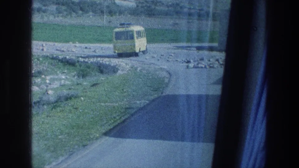 1976 8mm travel view near Jerusalem showing a yellow van on an unpaved road with grass, rocks, and fields nearby.