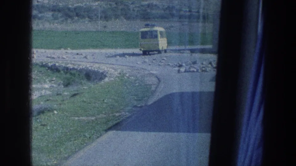 1976 Jerusalem-area home-movie frame with the rear of a yellow bus on a rural road near open grass and stones.