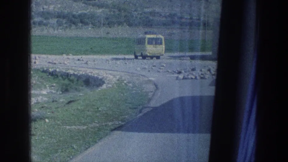 1976 Jerusalem-area view of a yellow bus traveling away on a curving rural road, partly framed by a dark window and curtain.