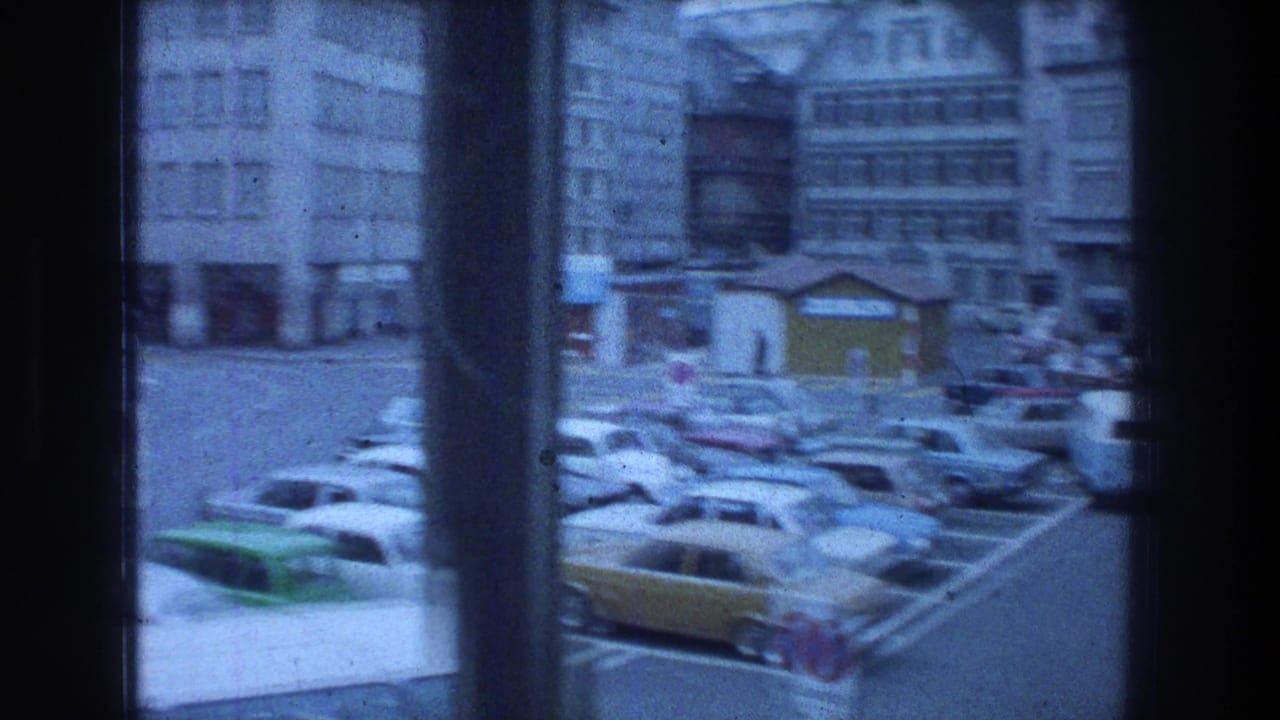 Zurich street seen through a window, with 1970s parked cars and multi-story city buildings beyond.