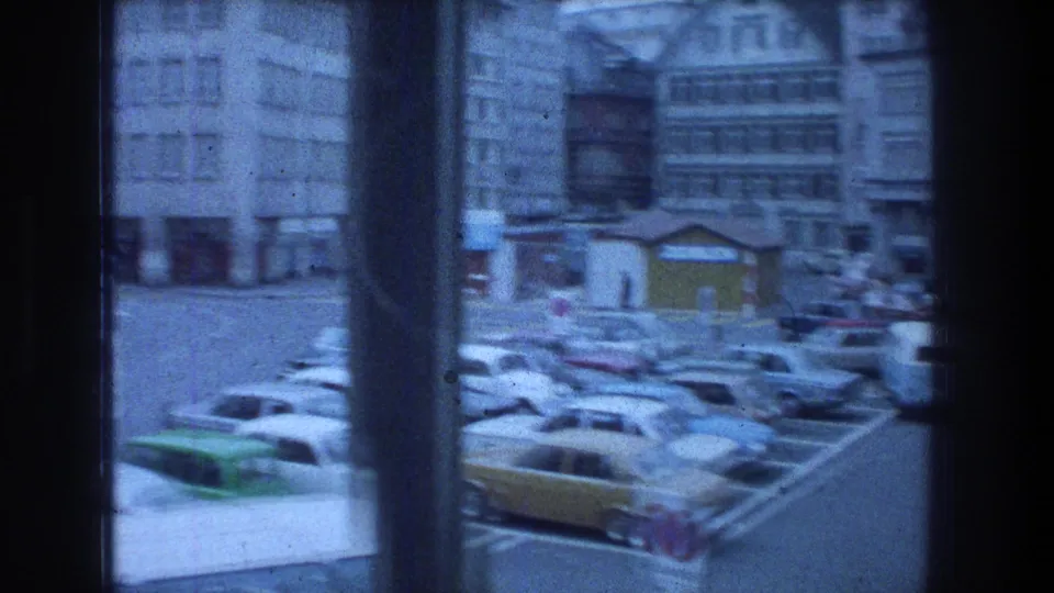 Zurich street seen through a window, with 1970s parked cars and multi-story city buildings beyond.
