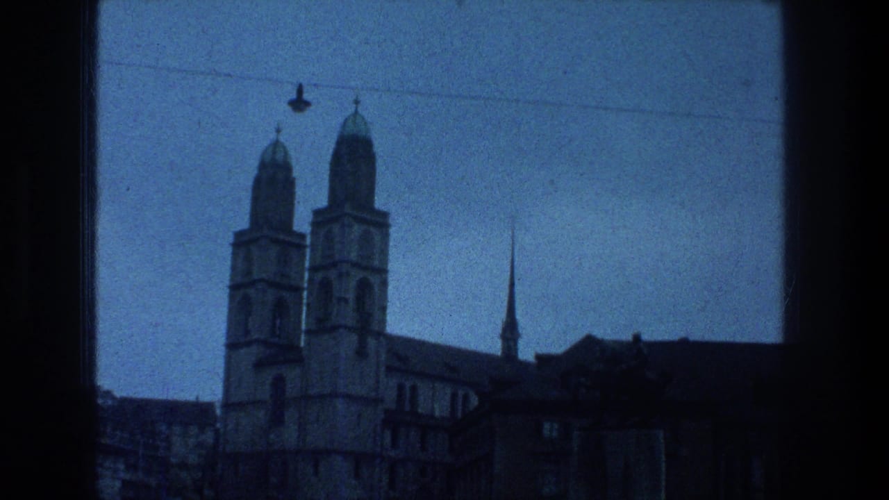 Zurich landmark view with twin church towers, rooftops, and a spire seen through a window-like frame on blue-tinted 8mm film.