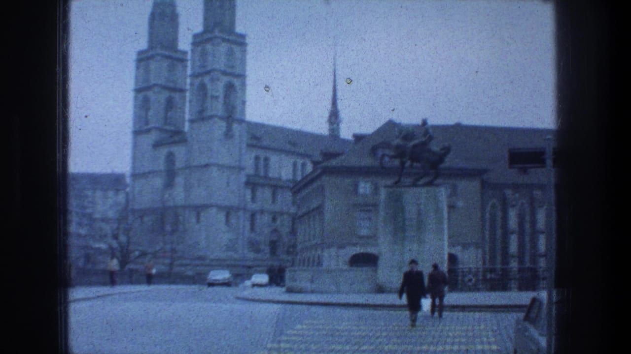 Twin church towers, square, cars, and two pedestrians walking near a statue.