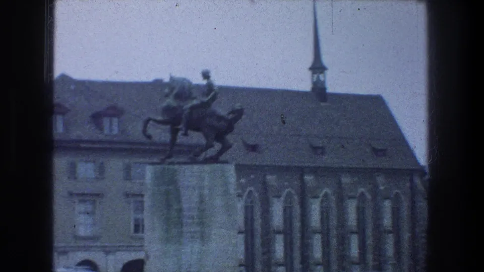 1976 Zurich home-movie frame showing an equestrian statue before older buildings and a church spire, with soft blue 8mm color.