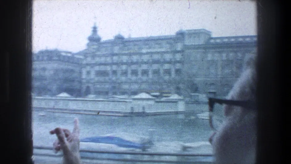 1976 Zurich tour view across water toward multi-story buildings, with a railing and a glasses-wearing passenger partially visible in the foreground.