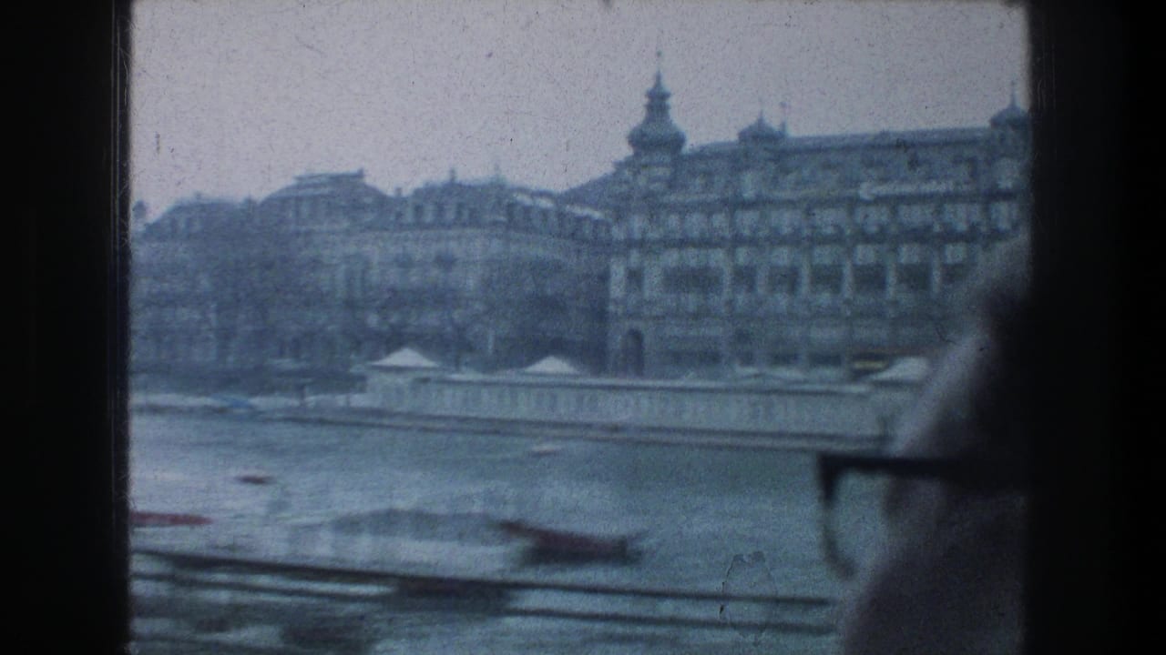 Zurich, 1976: a soft blue 8mm frame looks across the river toward historic buildings, with a partial passenger profile at the right edge.