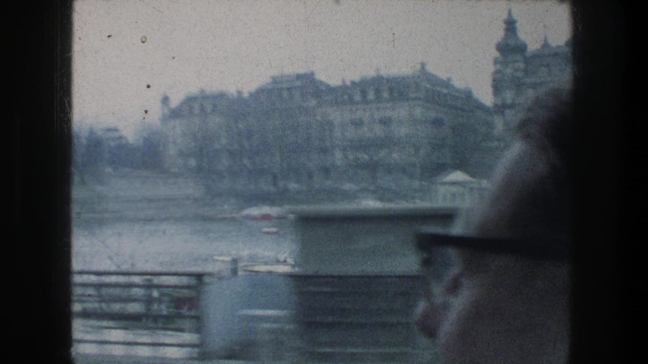A man in glasses sits by a train window as 1976 Zurich riverfront buildings, railings and boats pass outside.