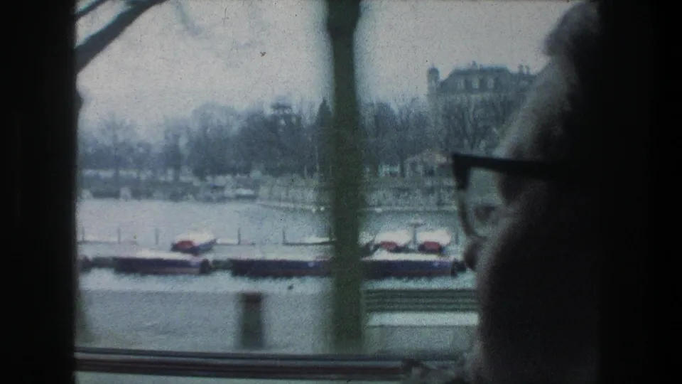 The Zurich lakeside view blurs slightly behind a person in glasses, with a dock, covered boats, bare trees, and railing visible through the window.