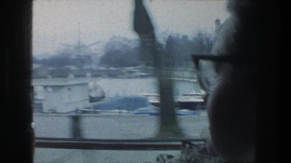 In Zurich, Switzerland, a woman in glasses sits by a moving vehicle window as boats and bare trees pass outside in 1976 home-movie footage.