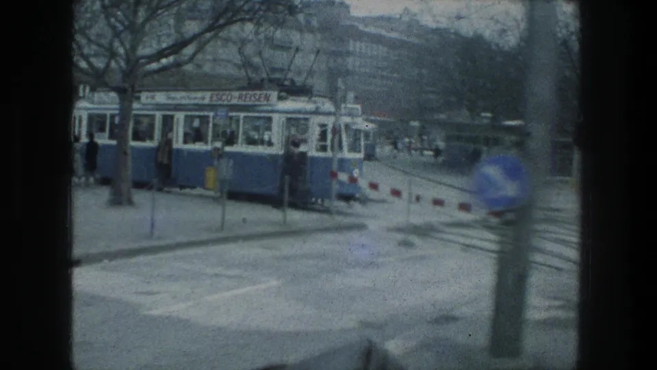 A blue and white trolley or tram with visible signage passes near a Zurich lakeside street, with trees, pedestrians, and barriers in view.