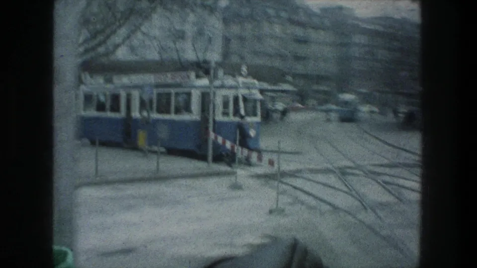 1976 Zurich street view from a moving vehicle, showing a blue-and-white tram, streetcar tracks, barriers, and city facades.
