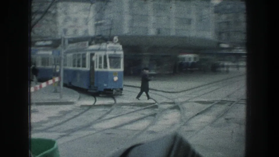 A pedestrian crosses the tracks near a blue-and-white tram in 1976 Zurich, with overhead wires and city buildings behind.