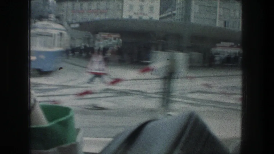 Motion-blurred 1976 Zurich street view with tram tracks, a partial tram, pedestrians, cars, and red-and-white barriers.