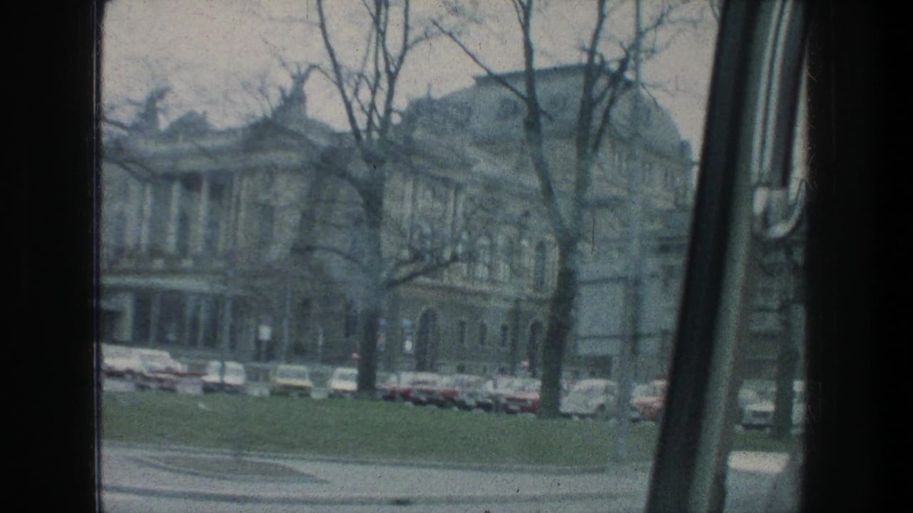 Zurich exterior in 1976 with a large multi-story building, bare trees, parked cars, and a vehicle window edge in the foreground.