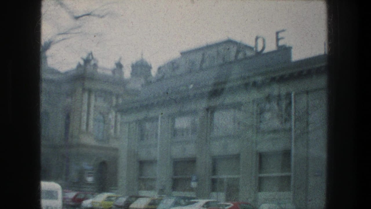 Soft 8mm frame from Zurich in 1976 showing a building facade, partial rooftop letters, parked cars, and the edge of a car window.