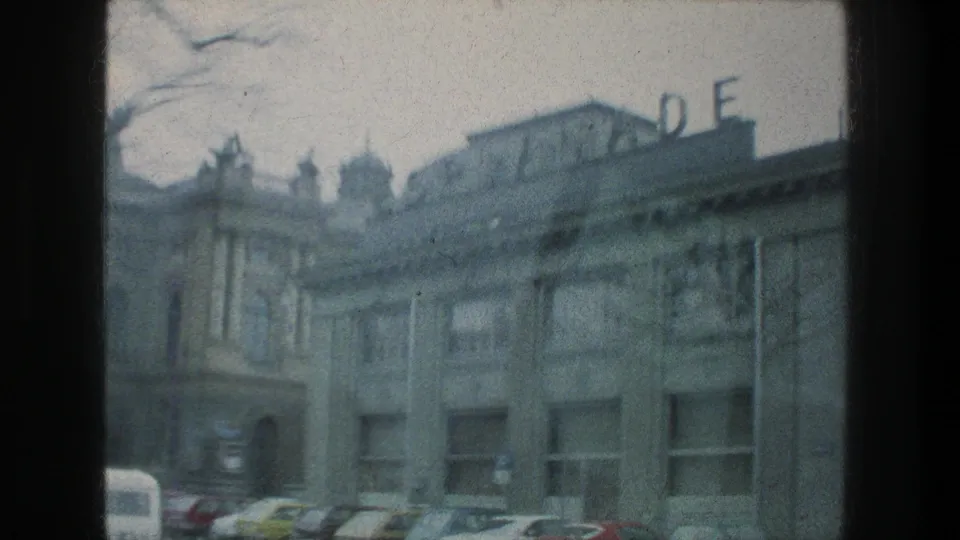 Soft 8mm frame from Zurich in 1976 showing a building facade, partial rooftop letters, parked cars, and the edge of a car window.