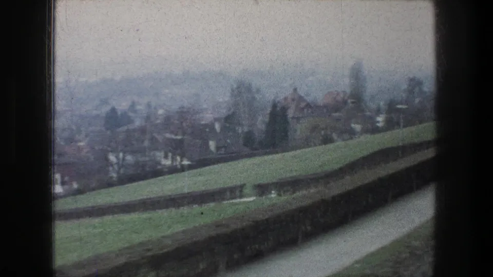 1976 Zurich-area bus-window view of a green slope, trimmed hedge, path, rooftops, and hazy trees.