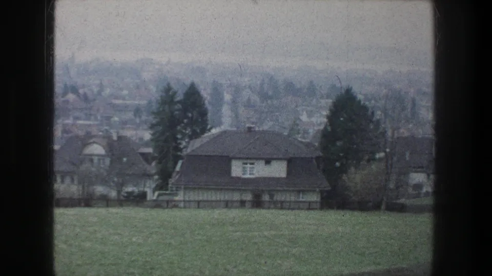 1976 Zurich home-movie frame showing a grassy slope, nearby houses, trees, and a soft distant neighborhood view.