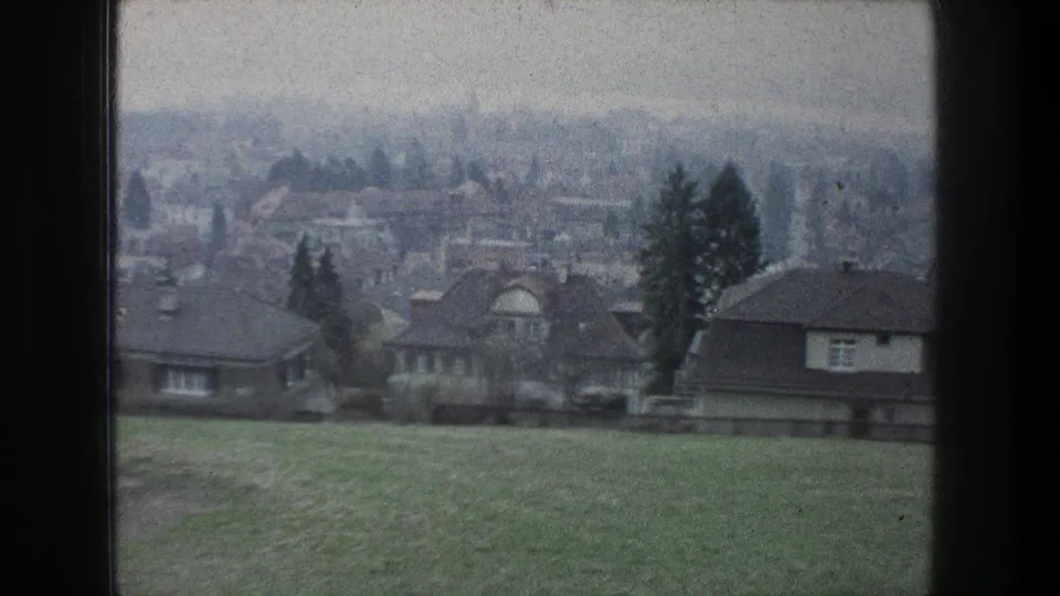Soft 8mm frame from Zurich in 1976 showing hillside grass, residential houses, rooftops, trees, and a hazy urban background.