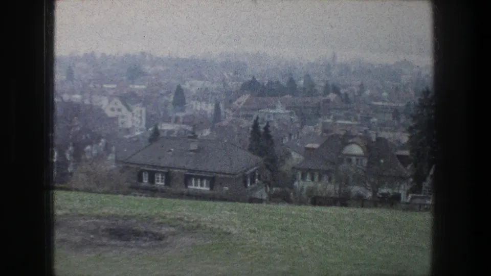 1976 Zurich home-movie frame showing a hazy town view from a grassy slope, with houses, bare trees, and an overcast sky.
