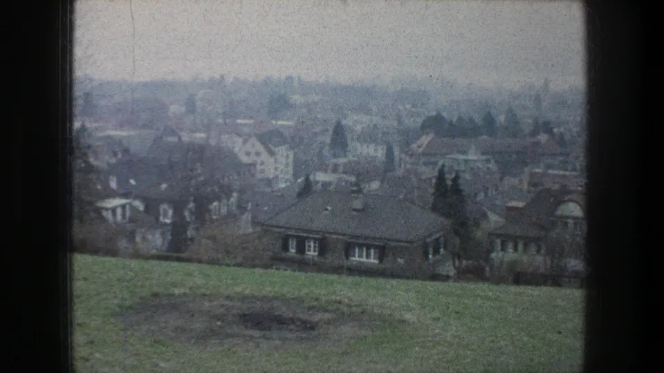 Hazy 1976 Zurich view across a grassy foreground toward houses, trees, and low buildings in muted 8mm color.