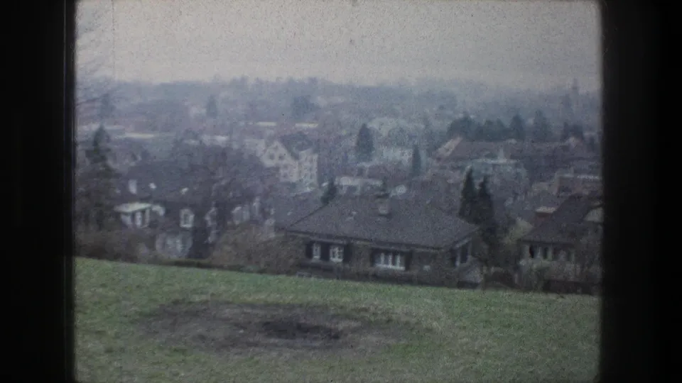 Zurich hillside view in bleached 8mm color, looking over grass toward houses and trees beneath a hazy sky.
