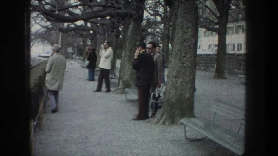 1976 Zurich park-path scene with several men in coats standing near bare trees, benches, and a low wall.